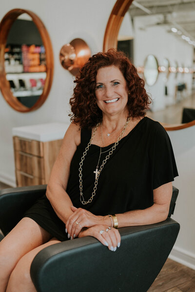 A professional headshot of Jauna French, the owner of Heath Salon and Spa. She is seated in a salon chair, smiling warmly with a confident and welcoming expression. The stylish salon background highlights her expertise and passion for the beauty industry.