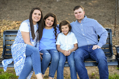 a mother, daughter, son, and father all dressed in matching blue sitting on a bench photographed by Millz Photography in Greenville, SC