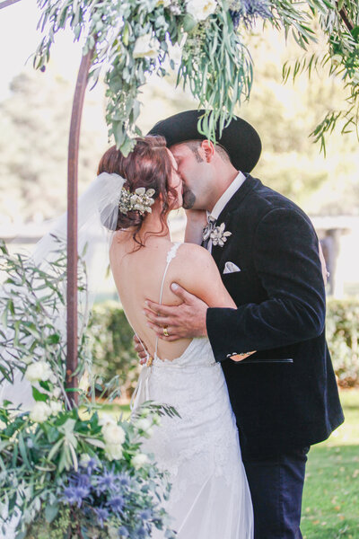 A groom in a cowboy hat kisses his bride under a floral arbor, blending rustic charm with wedding elegance.