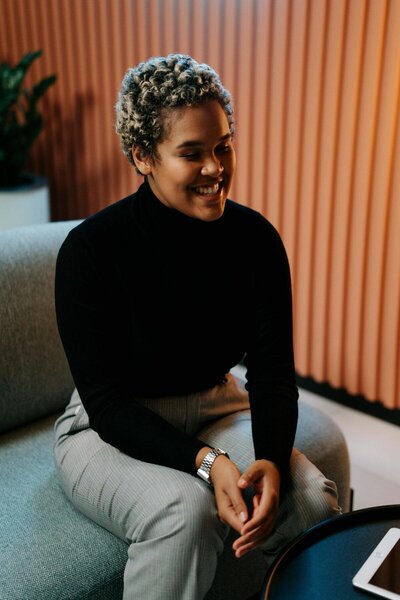 A person with short curly hair sits on a gray couch wearing a black turtleneck and gray pants. They are smiling and have their hands clasped in their lap. The background features a wooden vertical slat wall and a potted plant.
