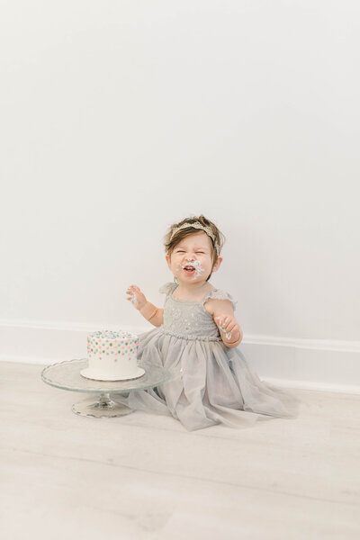 Smiling baby girl in a gray dress covered in frosting, sitting beside her birthday cake during a Kansas City first birthday milestone session.
