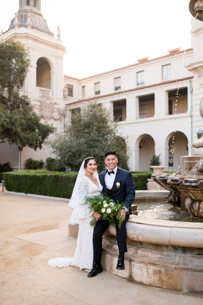 Groom sitting on the side of a fountain while a bride stands with her flowers next time in the courtyard at The City Hall in Pasadena, Caifornia.