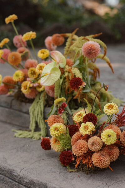 A lush, colorful bouquet is the main focus of the wedding photo with a couple embracing  partially featured. Part of the bride's face is showing, and the groom's black suit jacket is in focus. The wedding flowers are orange, yellow, and light blue.
