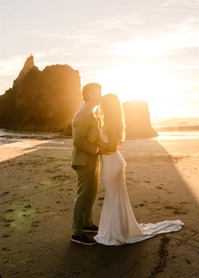 During the sunset of their Oregon Coast elopement, a couple stands backlit on a rocky beach. The groom kisses the brides forehead.
