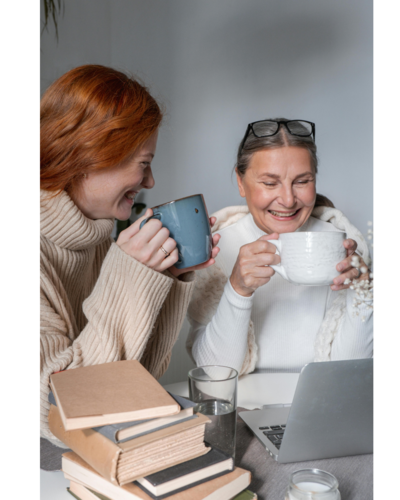 Two women with mugs laughing together in front of a laptop and books