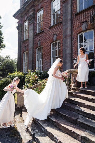 bride and bridesmaids all holding bouquets as they walk up stairs at their Shropshire wedding venue - the bridesmaids are in front and behind as they help the bride by lifting her dress up