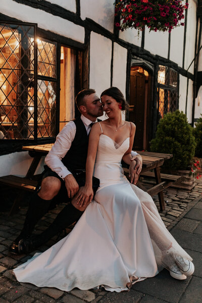 A bride and groom sit on a pub bench and embrace outside King's Chapel Amersham