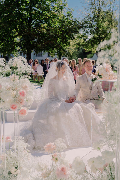A bride and a groom's  luxury lakeside wedding ceremony in Munich with a floating floral backdrop by the lake. The bride wears an Elie Saab bridal dress, holding a bouquet.