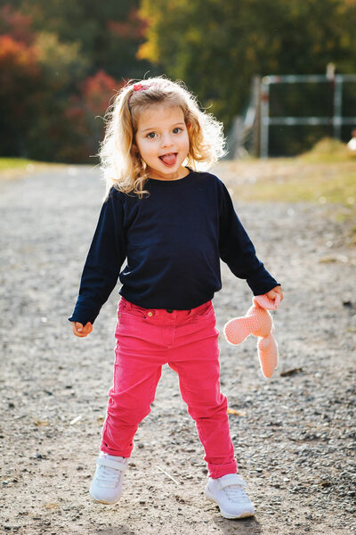 3 year old girl stands on a dirt path with the sun behind her. Her tongue is sticking out and she's holding a knit pink bunny.