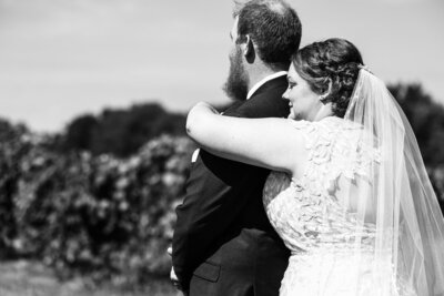 Bride wraps arm around groom's shoulder in an embrace among the grapevines at Lakeshore Vineyard & Events