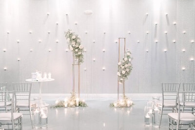 A modern wedding ceremony setup at SoHo63, a Chandler wedding and event venue, featuring two tall gold stands adorned with white florals and soft greenery. Dozens of floating candles and hanging lights create a romantic, minimalist backdrop.