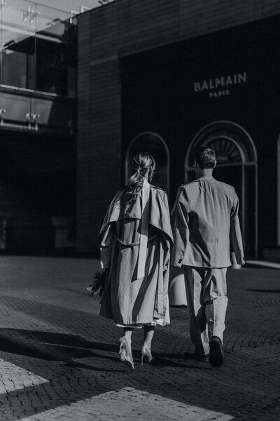 Black and white image of a woman and man walking towards a Balmain Paris store. The woman wears a white dress with a trench coat draped over her shoulders and stilettos, while the man dons a grey suit with his jacket casually slung over his shoulders.