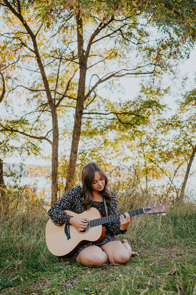 Young woman sits on grass playing guitar by Coeur d'Alene Lake