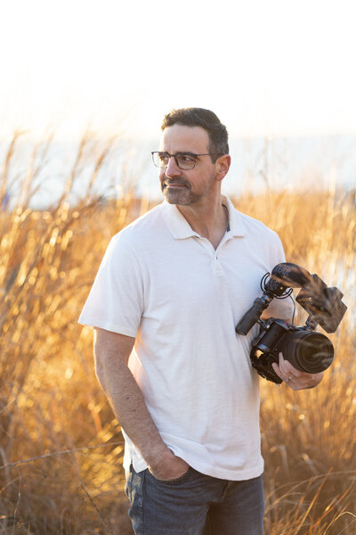 Brian Sellers, a destination wedding videographer based in Williamsburg, VA, looking into the distance holding a camera he uses to craft cinematic wedding films for brides and wedding couples