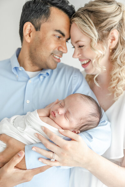 New parents smile forehead to forehead while holding sleeping newborn baby during baby photography session