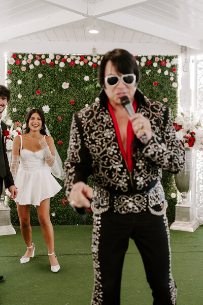 An Elvis impersonator singing with a newlywed couple laughing behind them 
