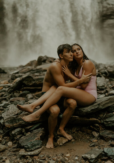 photo of couple holding each other in their arms, sitting in front of a water fall and look up to the right corner of the image. The man is in a bathing suit and shirtless, and the girl is in a one piece bathing suit.