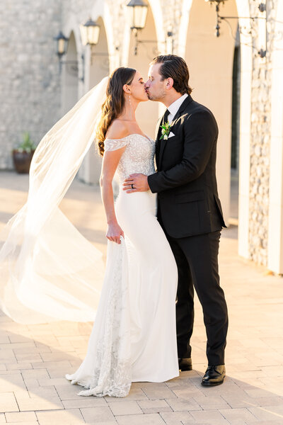 Bride and groom share a sunset kiss outside a stone and stucco venue, with the bride’s long veil blowing in the breeze and her off-the-shoulder gown catching the golden light. Captured in Westlake, Texas.