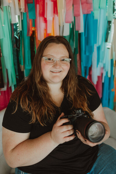portrait of mara shields, owner of M20 Media, sitting and smiling, with her camera, gimbal, balloons, and a hot pink background