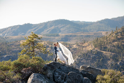 A couple stand on top of a large granite boulder in Yosemite on their elopement day and the groom holds the bride in the air as her veil flows behind her.