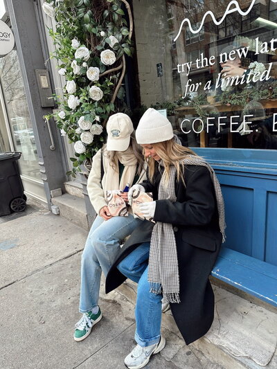 Two people sit on a blue bench outside a cafe, holding coffee cups. They wear winter outfits with beanies and scarves. White flowers decorate the window.