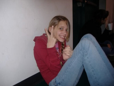 Girl in jean and a red hoodie sits against well while cheesing and holding up a peace sign.
