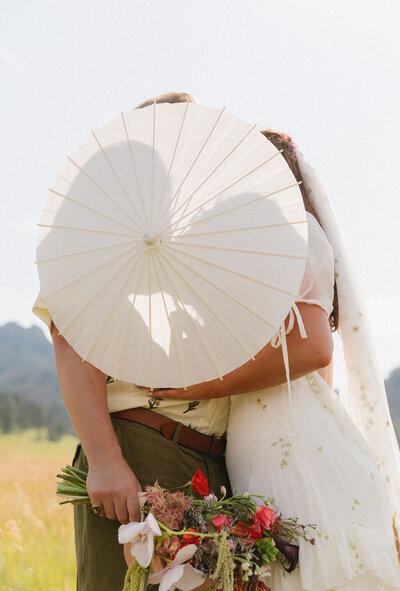Bride and groom hiding playfully behind a white parasol with bouquet visible in a Colorado meadow – whimsical Colorado wedding photos