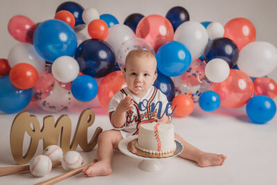 Atlanta, Georgia cake smash photographer photographing one year old boy sitting down with baseball smash cake in front of him on cake stand and red, white and blue balloons behind him