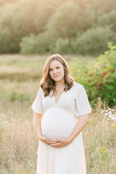 Pregnant Mom in a cream dress with shoulder length brown hair holding her baby bump. She is looking at the camera with a soft smile on her face and standing in a golden grassy field at sunset.