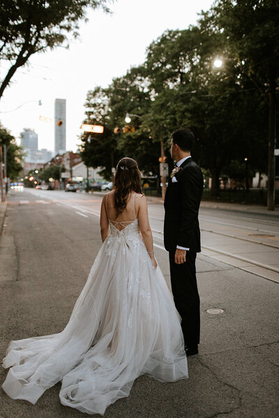 A couple in formal attire stands in an empty street at dusk, facing away. The woman is in a white gown, and the man is in a black suit. As trees and buildings line the street, Toronto wedding photographer Jennifer van Son captures a serene moment that embodies elegance and timeless romance.