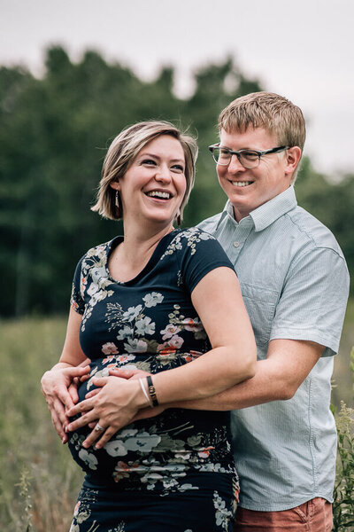 A couple expecting their second child laughs together with hands on her belly during a pregnancy photoshoot in Minnesota.