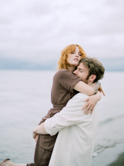 A man lifts his redheaded partner in his arms as she parts her lips in rapture on the shore of Presque Isle in Lake Erie. | Family Photographer in Pittsburgh PA | Anna Laero Photography