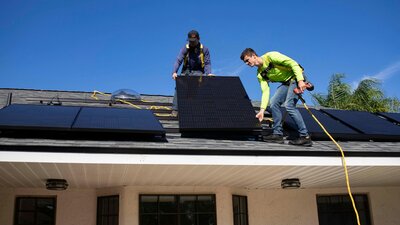 Two solar technicians installing black solar panels on a residential roof under a clear blue sky
