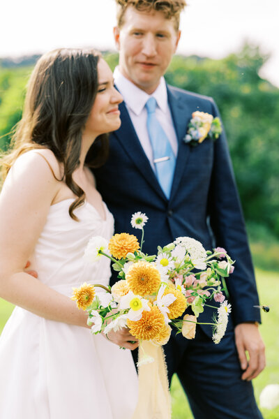 The groom looks upon his smiling bride holding a bouquet of flowers by Moonlight Floral Co, captured on film by photographer My Sun and Stars.