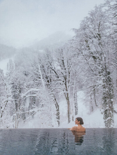 "A woman relaxes in a steaming outdoor infinity pool surrounded by snow-covered mountains, creating a serene contrast between warmth and winter’s chill.