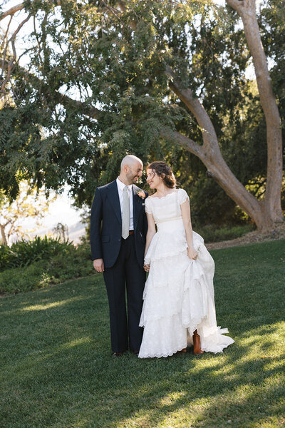 0-COVER-radiant-love-event-groom-holding-bride-looking-at-eachother-in-white-arched-outdoor-corridor-romantic-elegant-timeless