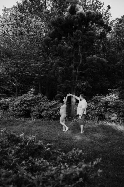Black and white maternity photo of mom and dad dancing in the distance in front of trees in a garden in Baltimore Maryland.