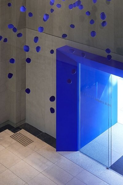 A lobby area featuring grey walls, marble flooring, a glass doorway surrounded by a deep blue frame, and a cluster of blue lighting fixtures hanging from the ceiling
