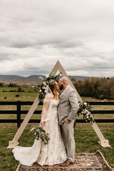 bride and groom kissing as they stand on a rug in the grass with a triangular wedding arch adorned with floral arrangements behind them at their Shenandoah wedding venue