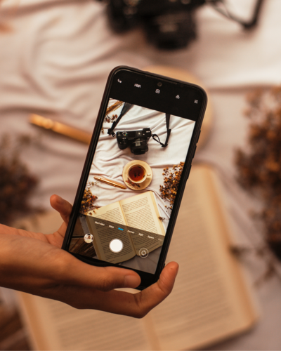 view of smart phone taking flatlay photo of a book, cup of tea, and camera