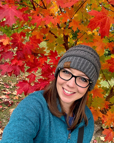 A woman standing in front of tree with fall colors of reds and oranges.  Woman wearing a gray stocking hat, black glasses, blue eyes, brunette shoulder length hair, and wearing a blue long sleeve jacket.