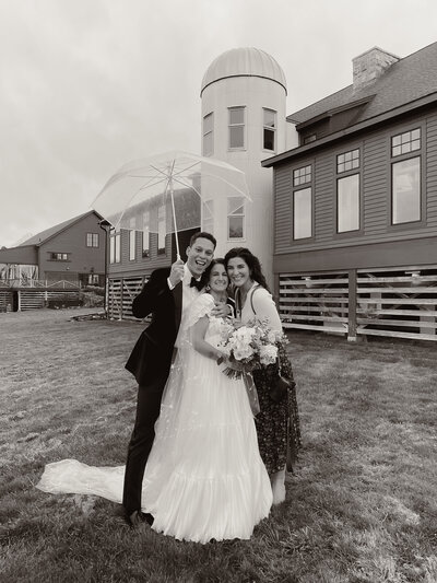 Bride and groom posing with wedding photographer under a clear umbrella