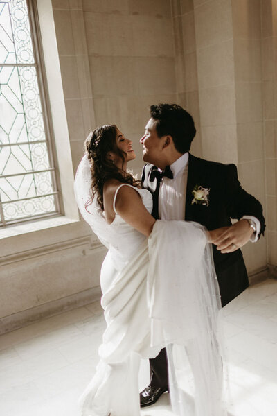 Bride in a white gown and groom in a black suit share a joyful dance by a large window with ornate patterns during their San Francisco City Hall wedding photoshoot, bathed in natural light.