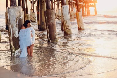 A serene sunrise casts a warm glow on a pregnant woman in a light blue dress standing in the water.