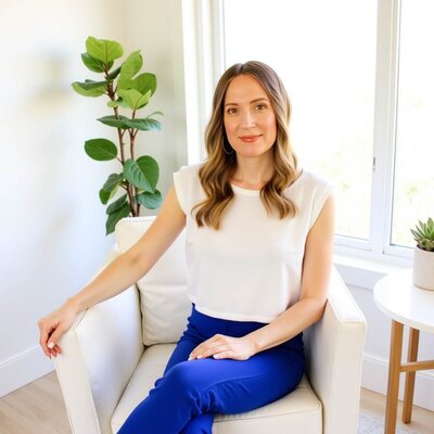 Stacey Xanthe from Xanthe Bookkeeping sitting in a white leather chair in a bright, cozy living room, wearing a white top and cobalt blue pants, with a side table, succulent, and green leafy plant in the background.
