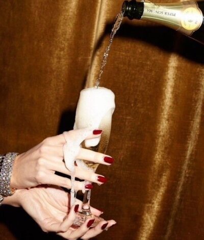 A woman with red painted nails holding a Champagne glass that is overflowing as someone overflows it