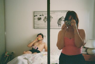 Woman taking a mirror selfie with a film camera while her partner lounges in bed in the background, capturing a candid romantic moment.