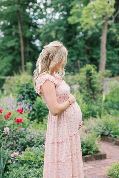 A pregnant woman wearing a pink floral  dress  stands in a garden holding her pregnant belly photographed by New jersey maternity photographer Kate Voda Photography