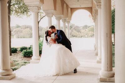 A groom dips and kisses his bride in front of stone arches.