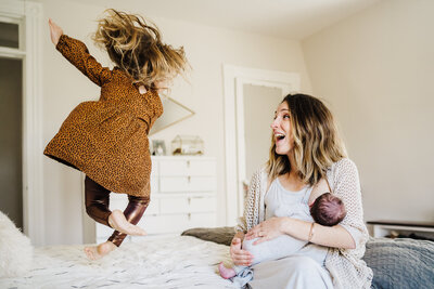 mother nursing newborn laughs as toddler jumps on bed